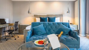 A hotel room with a large blue sofa, a glass table with a plate of fruit and champagne, and a desk with a computer.