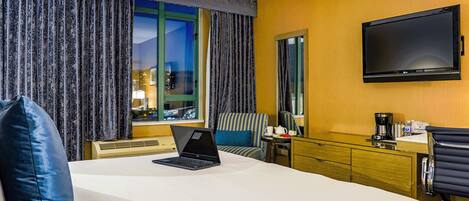 A hotel room with a bed, a desk with a laptop, a coffee maker, and a TV on the wall.