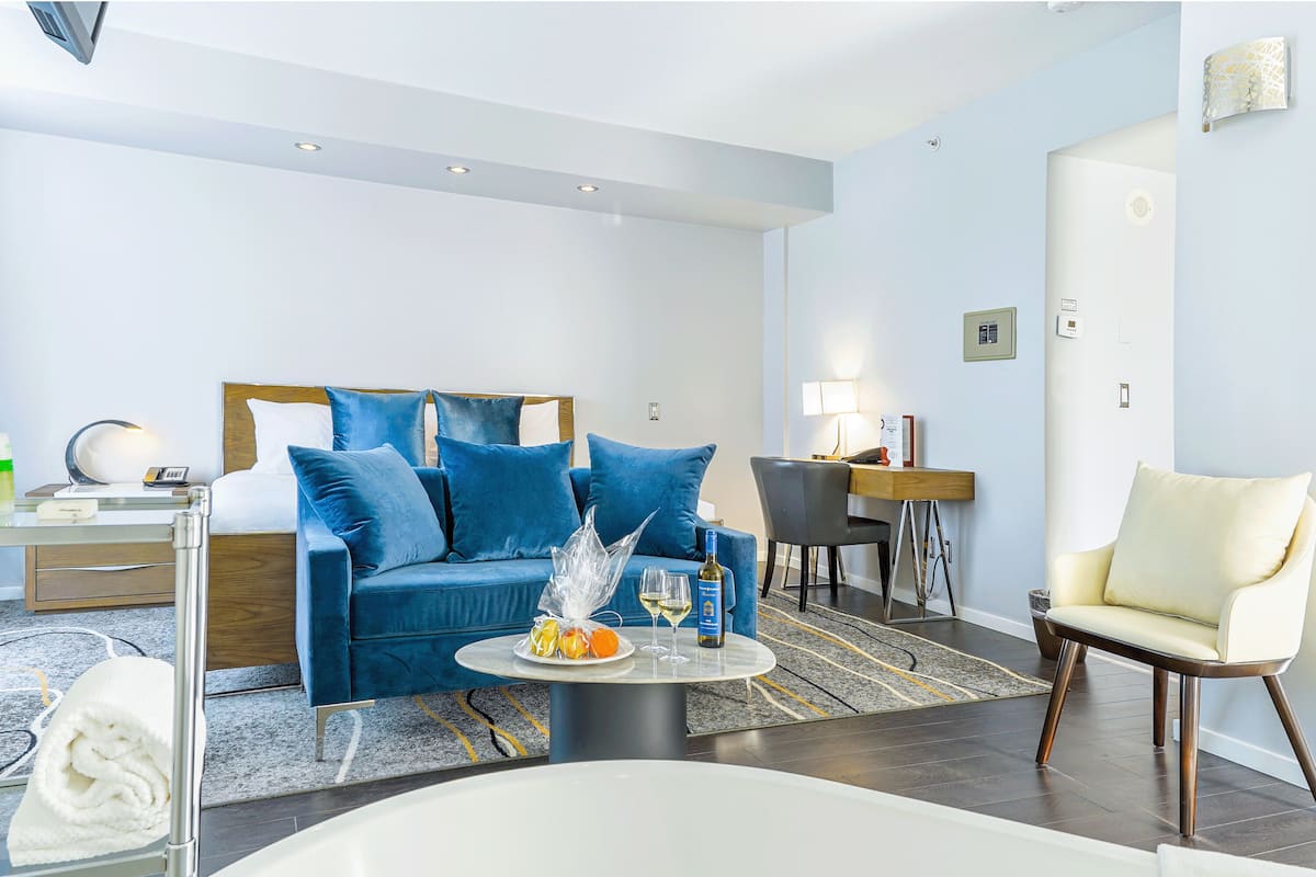 Ett modernt hotellrum med en blå soffa, en vit fåtölj, ett runt soffbord med en flaska och glas, och ett litet skrivbord med en lampa.