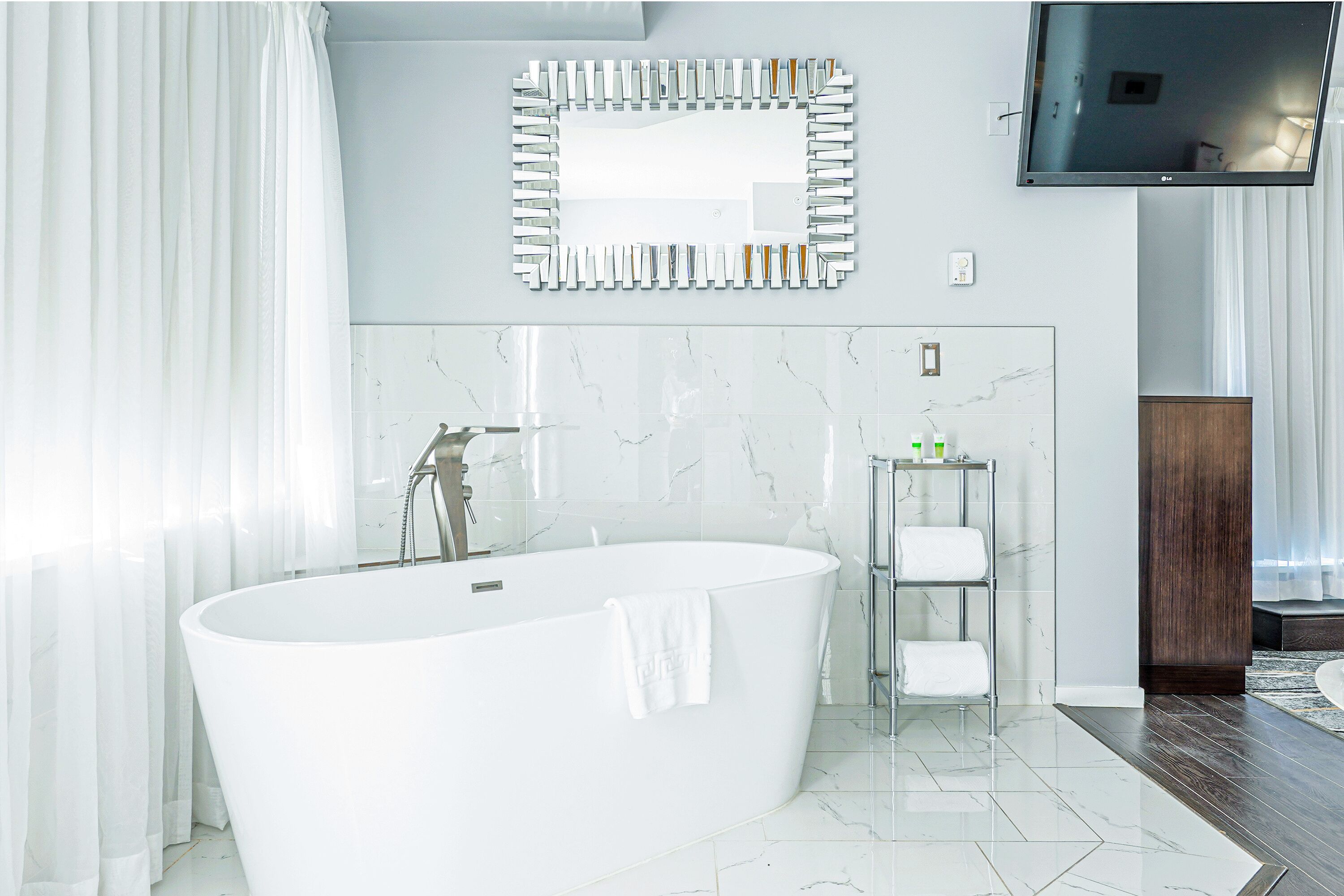 A modern bathroom with a freestanding bathtub, a mirror, a television, and a small shelf with toiletries.