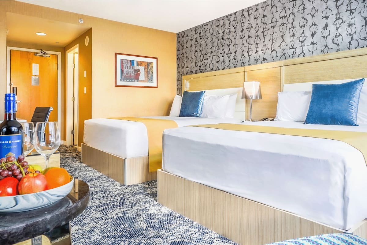Ett hotellrum med två sängar, en sänggavel i trä, en inramad tavla på väggen och ett bord med en flaska vin och frukt.