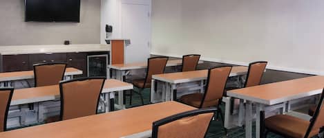 Un salón de conferencias con varias mesas y sillas, un televisor de pantalla plana montado en la pared y una puerta con un teléfono en la pared.