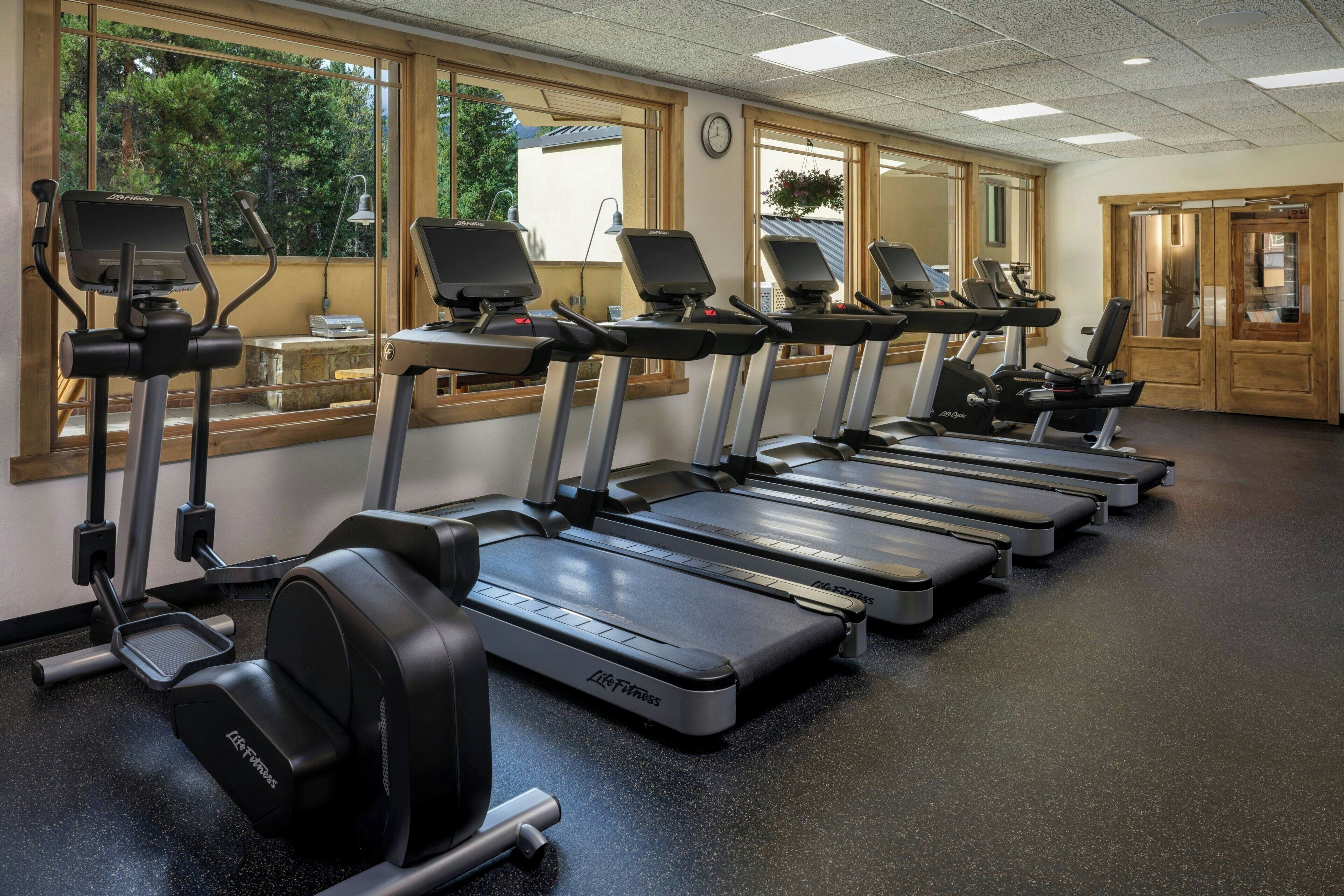 A gym with multiple treadmills aligned in a row, large windows with a view of greenery, and a wooden door.