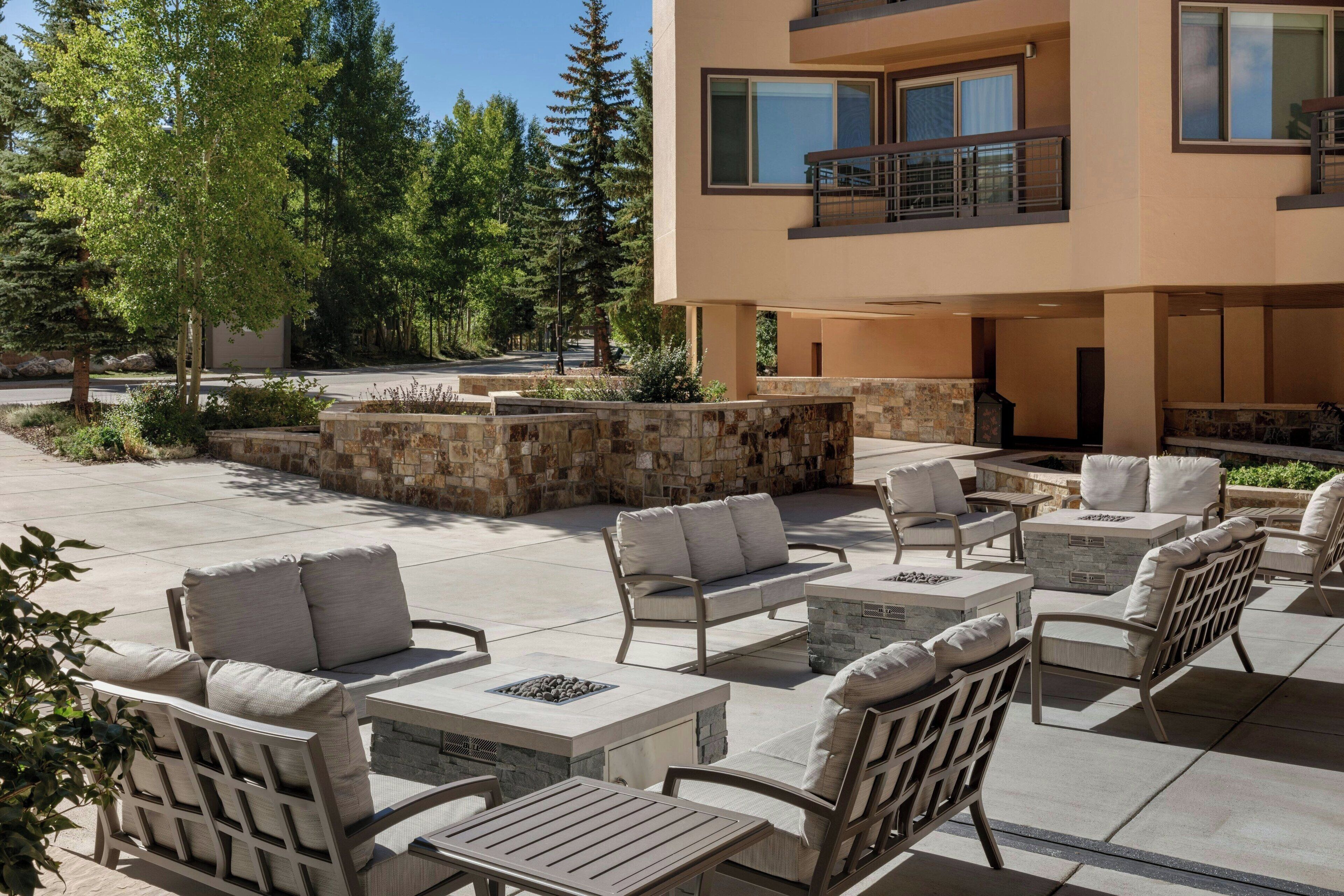 A modern outdoor patio with stone seating and a fire pit, surrounded by a stone wall and trees.