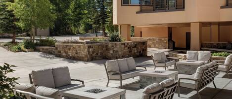 A modern outdoor patio with stone seating and a fire pit, surrounded by a stone wall and trees.