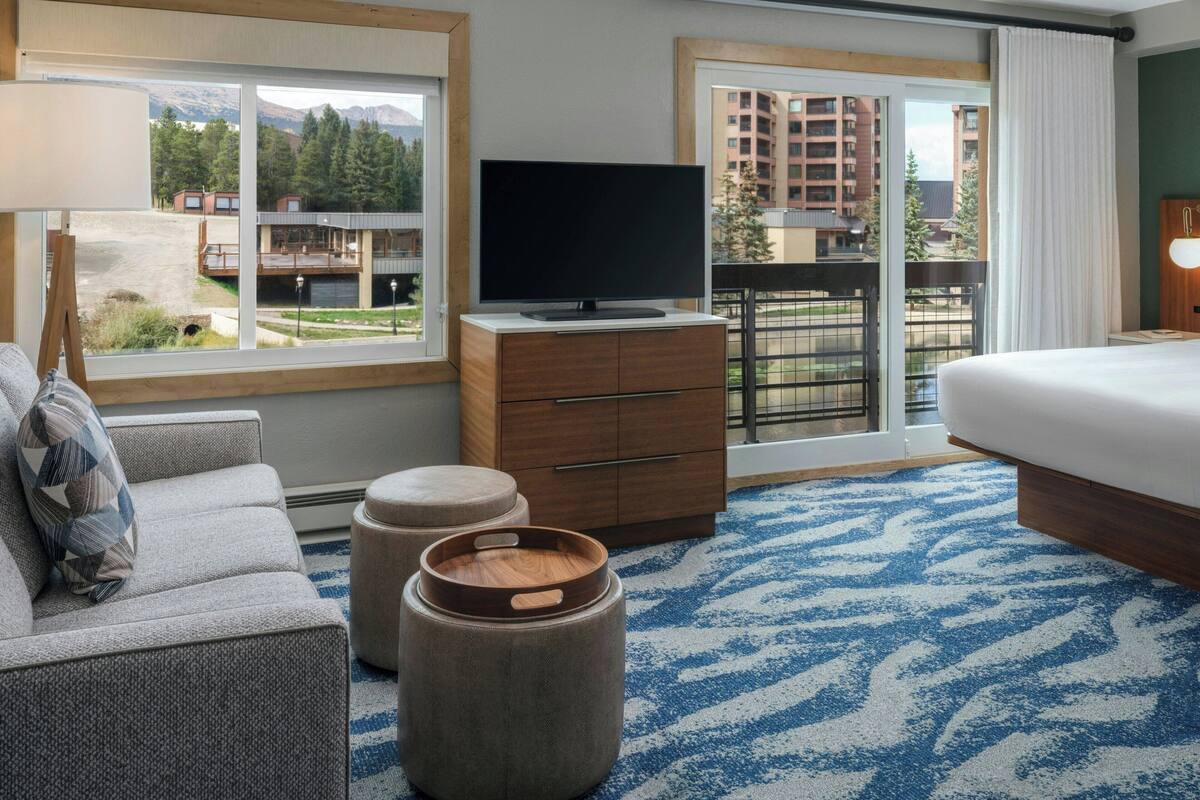 Habitación de hotel con una cama grande, un sofá, un televisor sobre un mueble de madera y vistas a un edificio y árboles a través de la ventana.
