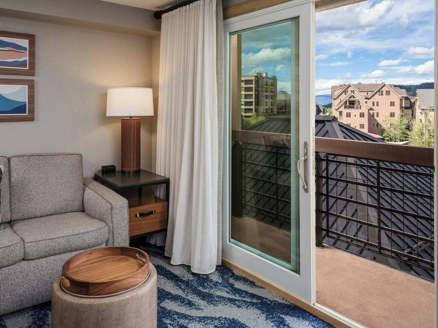 A hotel room with a sofa, a lamp, a small wooden side table, a round ottoman, and a sliding glass door leading to a balcony with a view of buildings and a cloudy sky.
