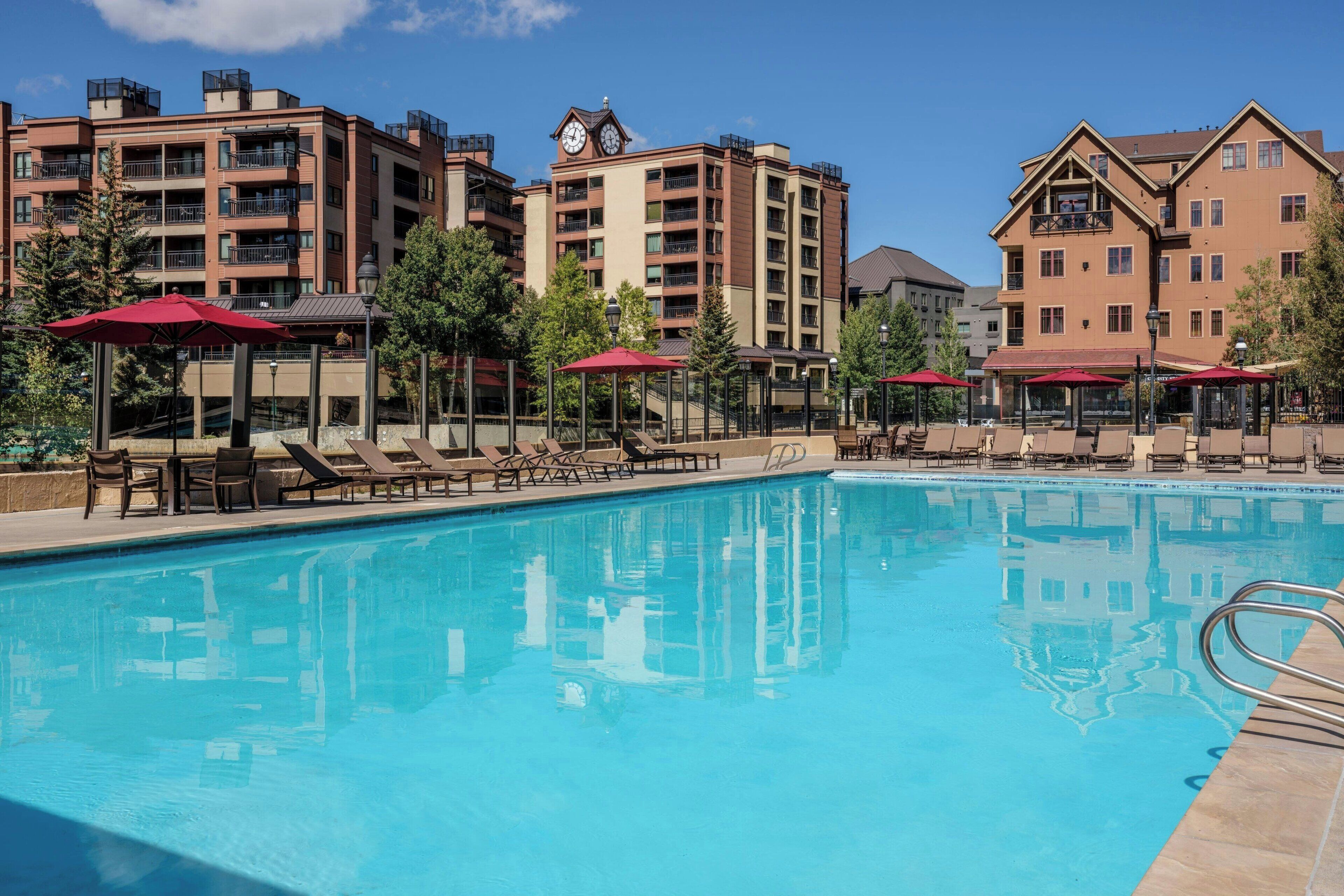 Una piscina con sillones de playa y sombrillas, rodeada de edificios de varios pisos con balcones y una torre del reloj.