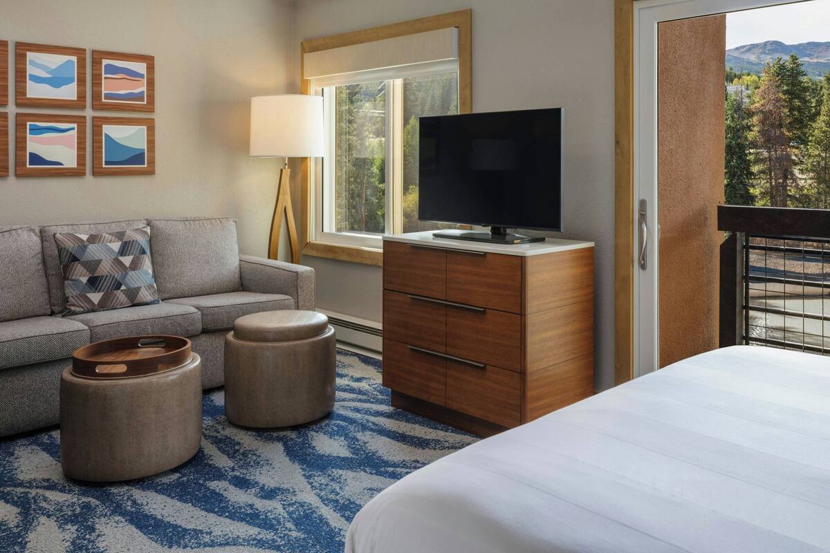 Habitación de hotel con una cama, un televisor sobre una cómoda de madera, un sofá, dos otomanas, una lámpara y una ventana con vista a árboles y montañas.