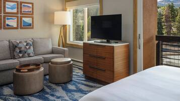 A hotel room with a bed, a TV on a wooden dresser, a sofa, two ottomans, a lamp, and a window with a view of trees and mountains.