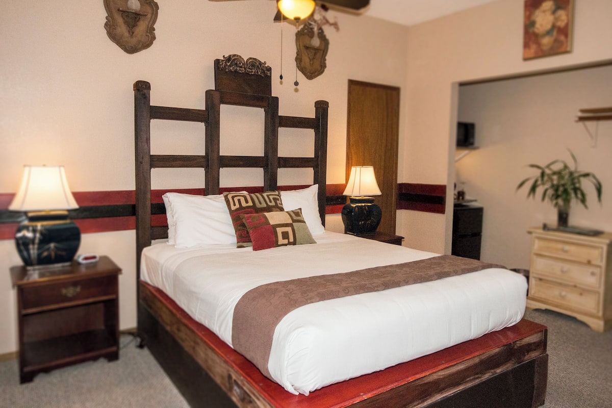 A neatly made bed with a wooden headboard, two bedside tables with lamps, a mirror, and a dresser with a plant.