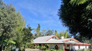 A house with a red roof and white walls, surrounded by lush greenery and trees. - Lokahi Lodge (Volcano)