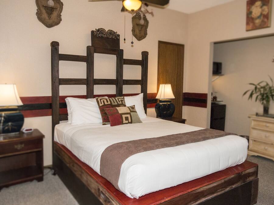 A neatly made bed with a wooden headboard, two bedside tables with lamps, a mirror, and a dresser with a plant.