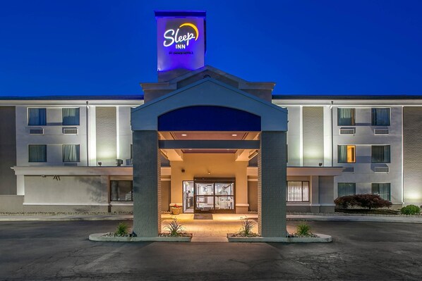 A Sleep Inn hotel entrance with a modern design, featuring a prominent sign, a covered entrance, and a landscaped area with plants.