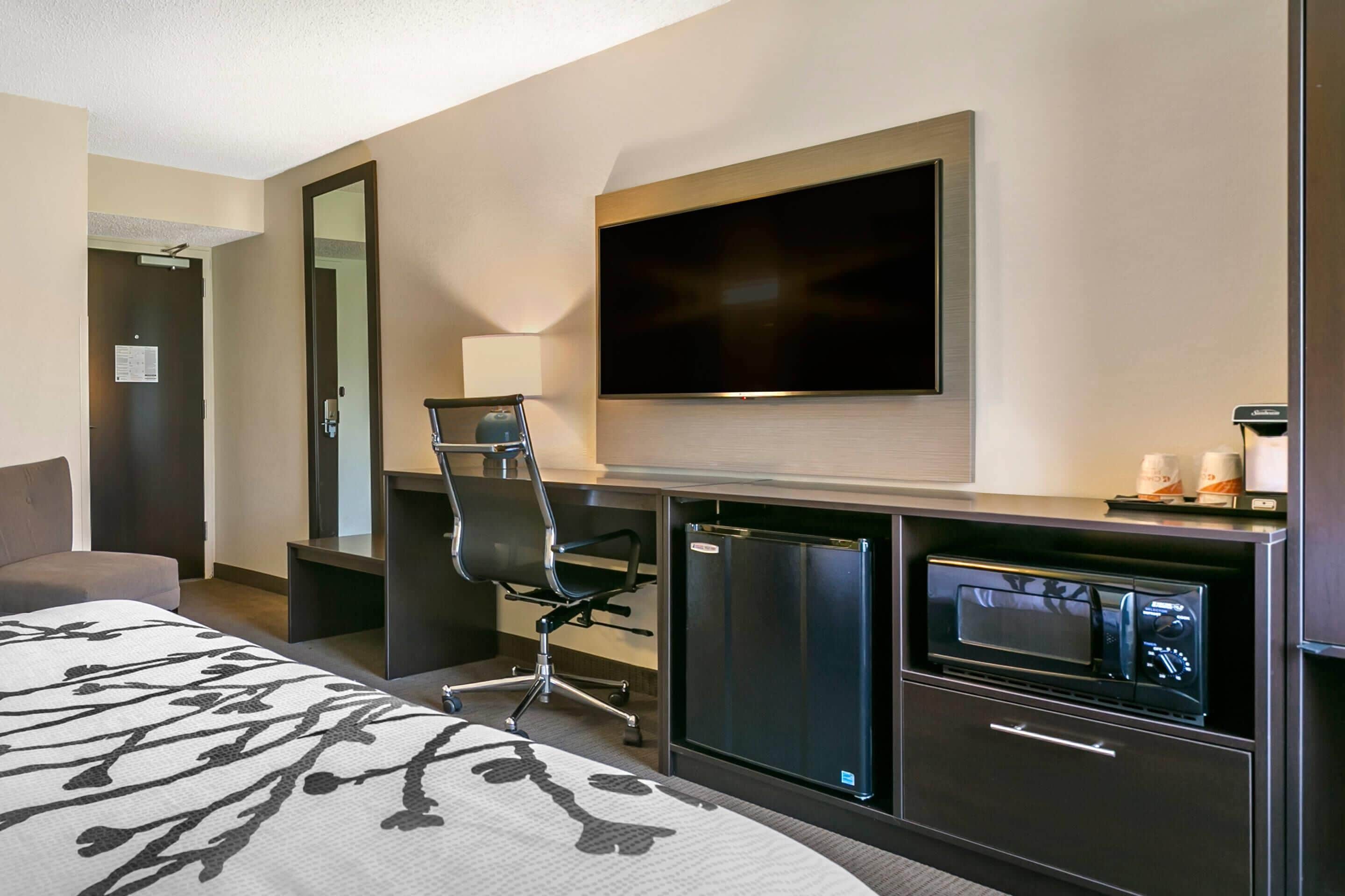 A hotel room with a bed, a desk with a chair, a flat-screen TV, a minibar, and a refrigerator.