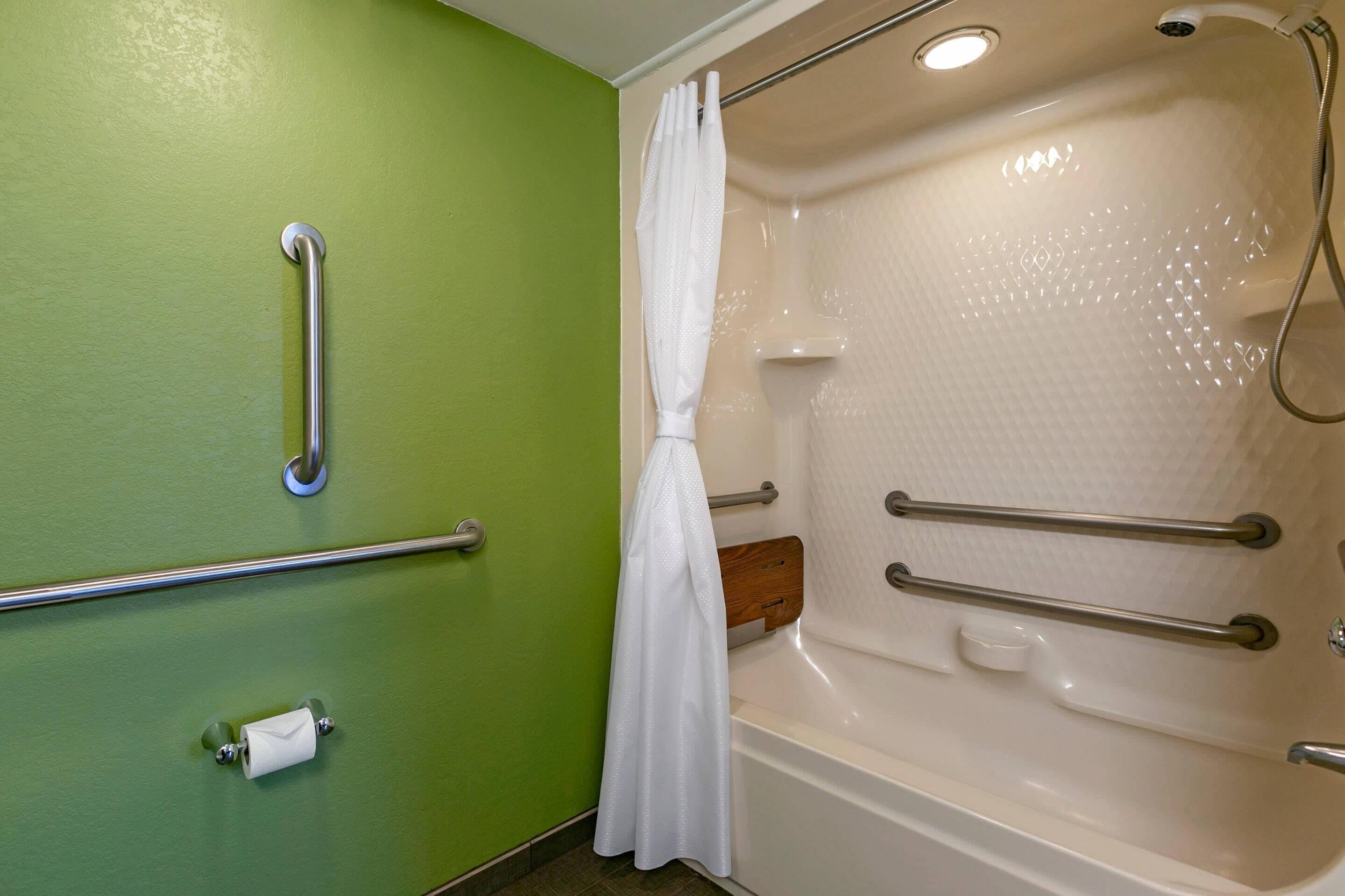 Un baño con paredes verdes, una bañera blanca con barras de apoyo, una cortina de ducha y un dispensador de papel higiénico.