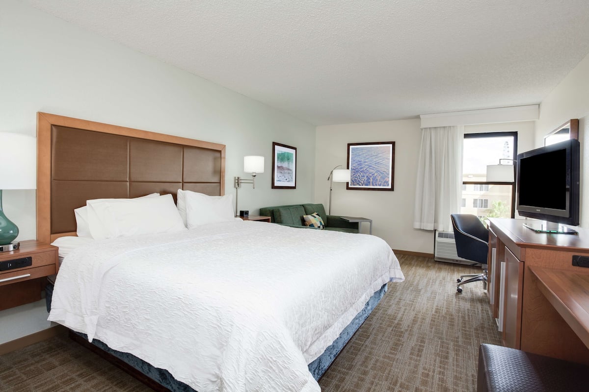 Une chambre d’hôtel avec un grand lit, un bureau avec une télévision, un fauteuil vert et une fenêtre avec des rideaux.