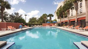 A swimming pool with lounge chairs and umbrellas, surrounded by palm trees and a building with red awnings.