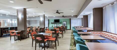 A spacious dining area with multiple tables and chairs, ceiling fans, and a television screen in the background.
