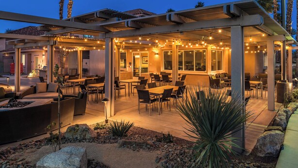 Un área de patio al aire libre con asientos y mesas, luces colgantes y una estructura de pérgola.