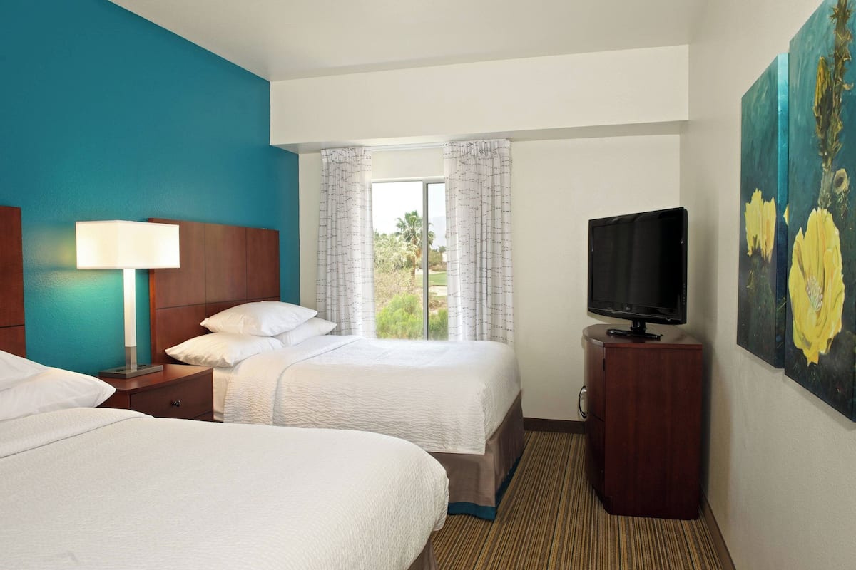 A hotel room with two beds, a TV, a window with curtains, and a painting on the wall.