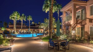 Zona de piscina con sillas de descanso y una mesa, rodeada de palmeras y un edificio con balcones.