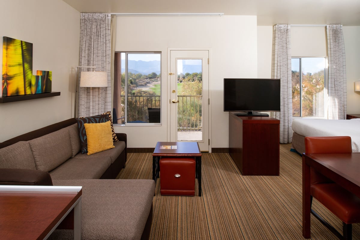 A hotel room with a sofa, a small table, a TV, and a balcony with a view of the outdoors.