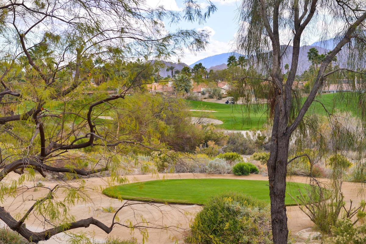 A golf course with sand bunkers, a putting green, and a backdrop of mountains and buildings.
