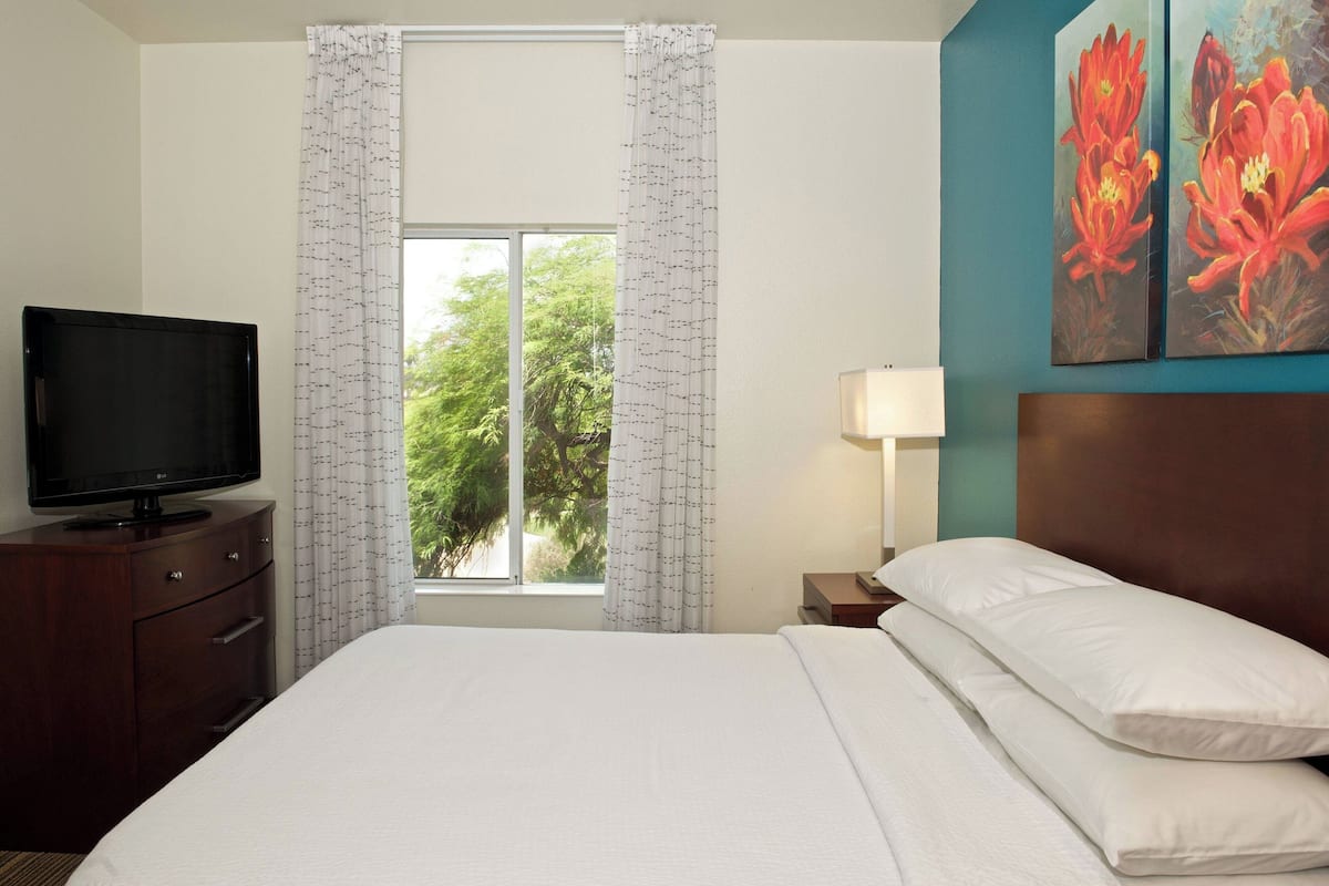 A hotel room with a bed, a TV on a wooden cabinet, a window with curtains, and a painting on the wall.