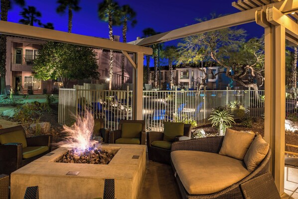 An outdoor patio area with wicker furniture, a fire pit, and a view of a pool and buildings in the background.