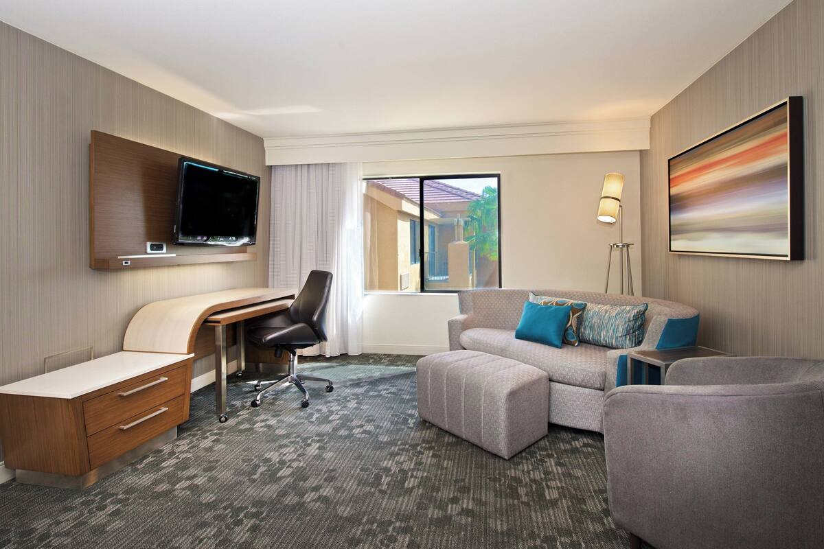 A hotel room with a desk, a sofa, a TV mounted on the wall, and a window with a view.