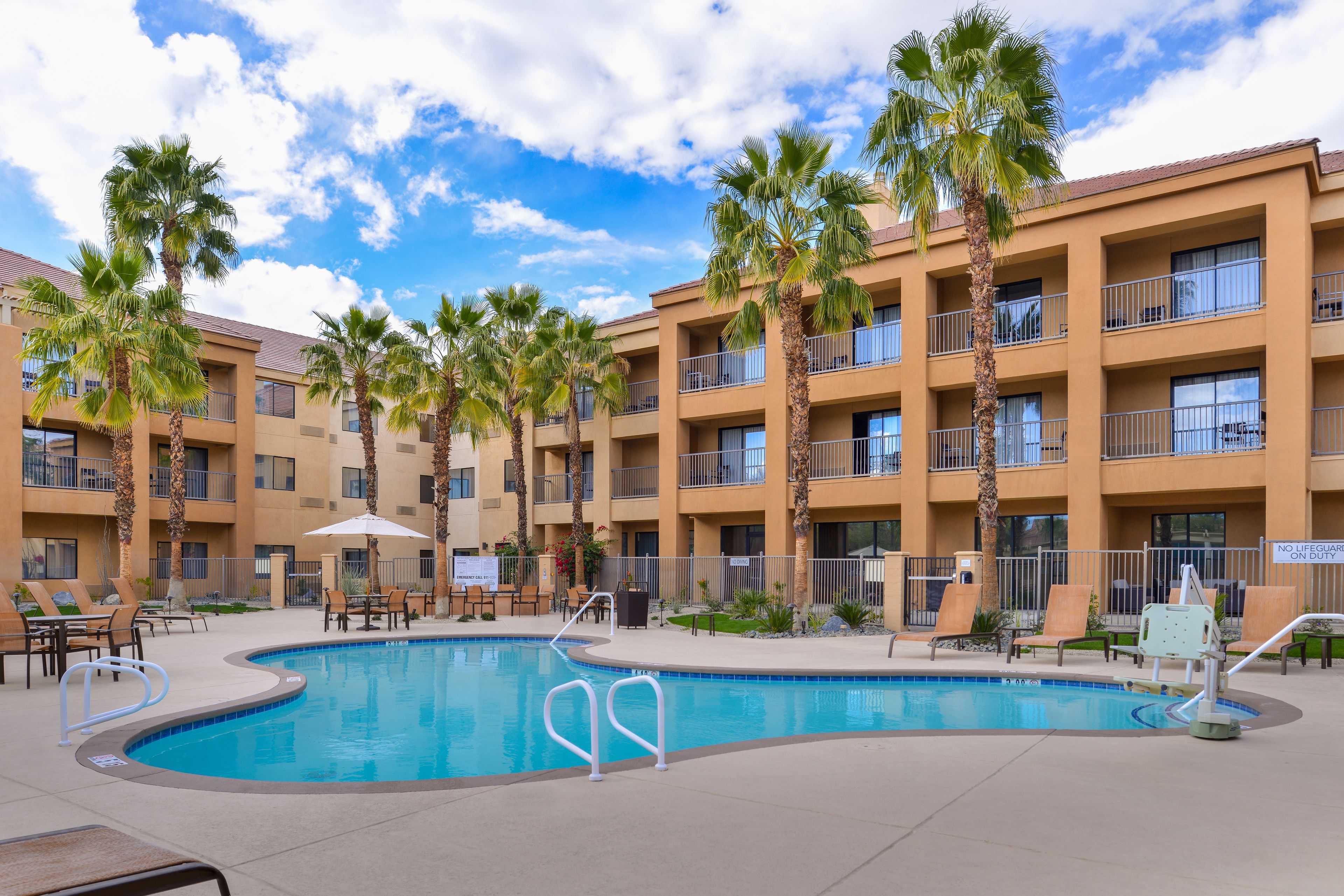 A pool area with lounge chairs and palm trees, surrounded by a multi-story building with balconies.