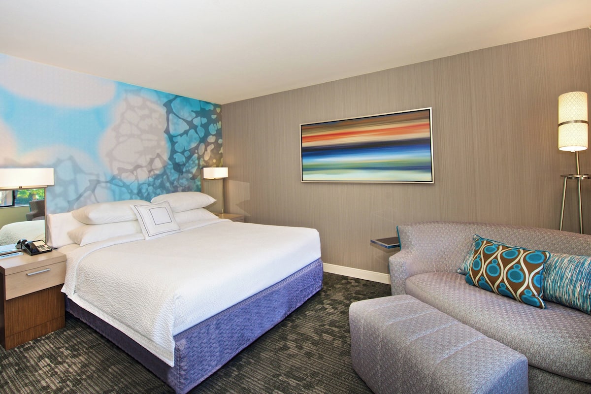 A hotel room with a large bed, a sofa, a desk with a lamp, and a painting on the wall.