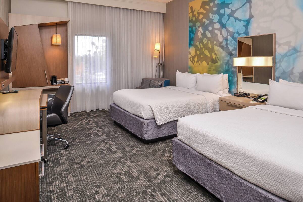 A hotel room with two beds, a desk, a chair, a TV, and a painting on the wall.