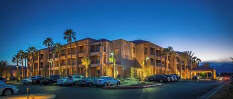 A multi-story hotel with a parking lot, palm trees, and a mountain in the background.