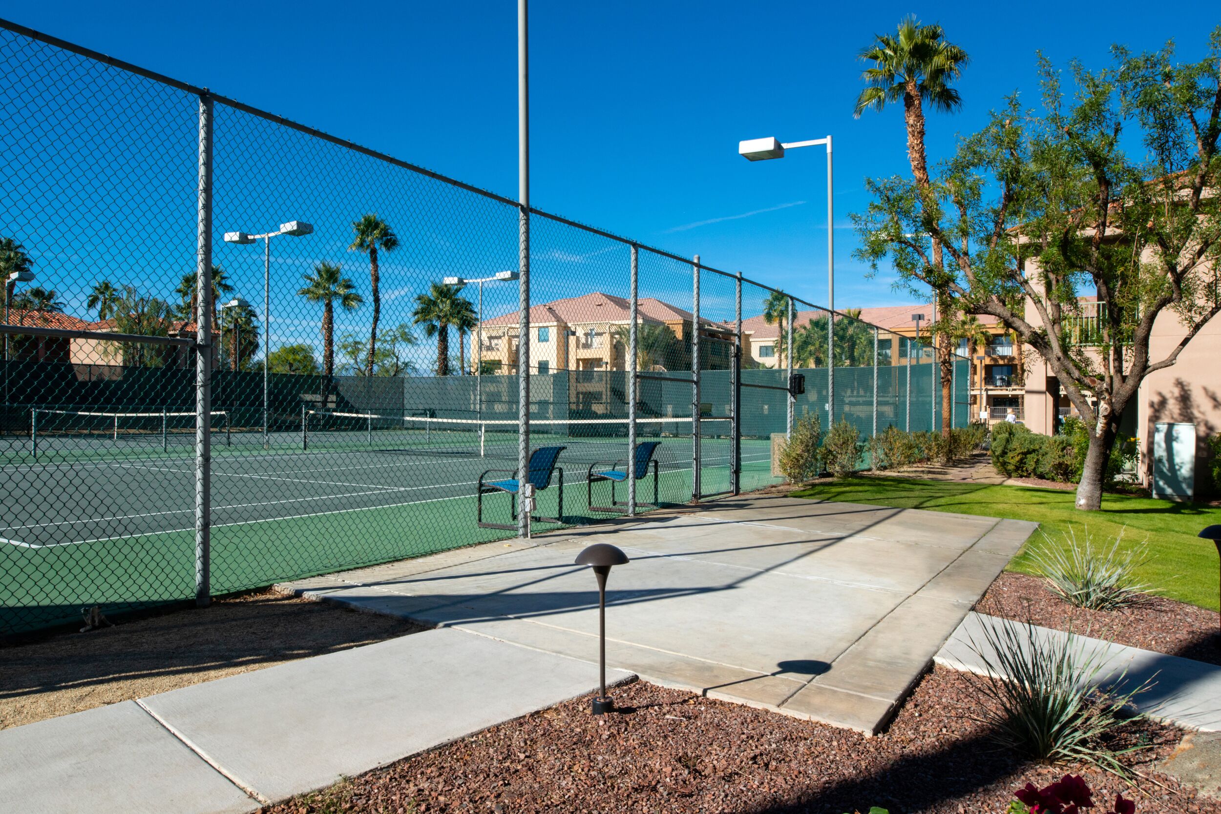 A tennis court with a chain-link fence, benches, and a street lamp.