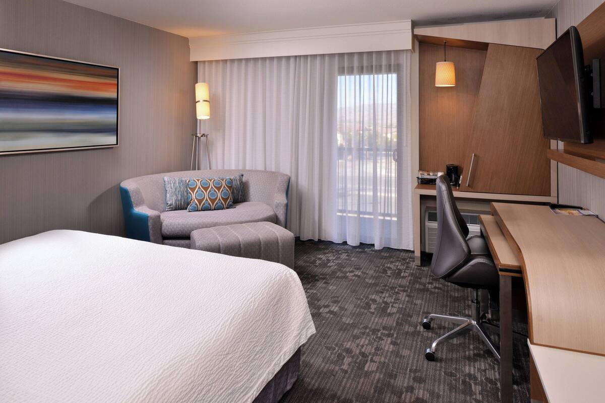 A hotel room with a bed, a desk, a chair, a TV, and a large window with curtains.