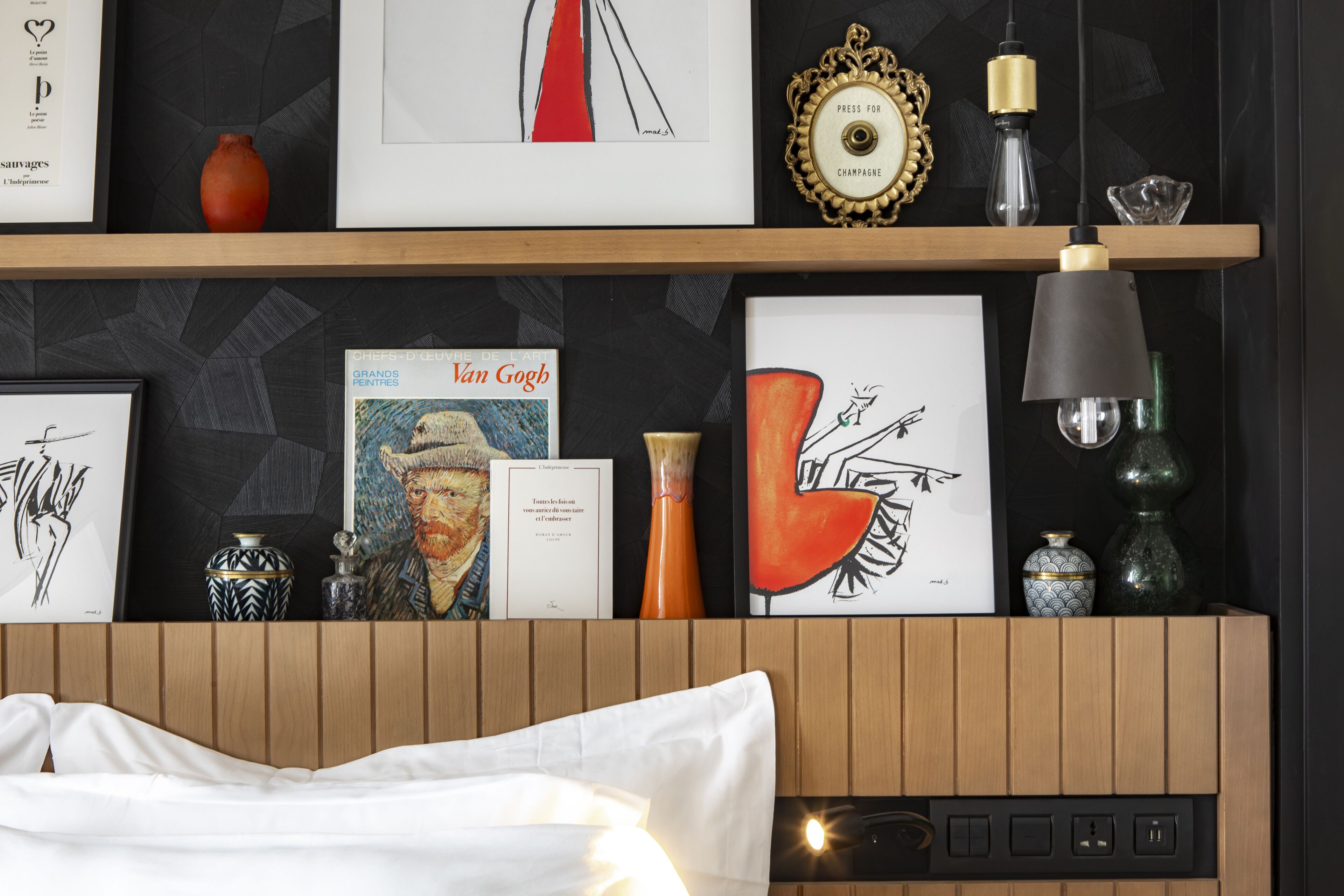 Una habitación de hotel con cabecera de madera, cama con ropa de cama blanca, estante de pared con varias obras de arte enmarcadas, un jarrón y una lámpara.