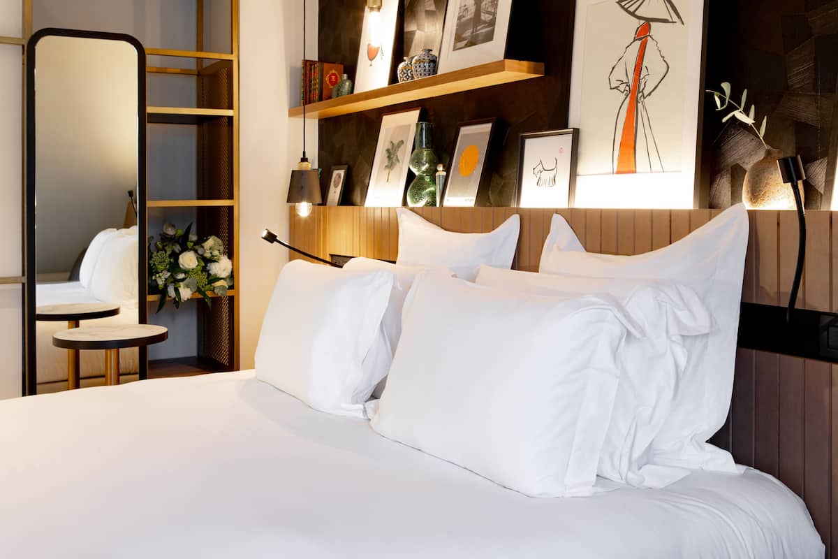 Una cama bien hecha con sábanas y almohadones blancos, una mesita de noche con una lámpara, un espejo y un estante con marcos y adornos.