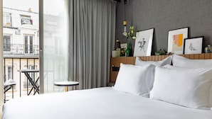 A hotel room with a large bed, a small round table, a balcony with a view of buildings, and framed artwork on the wall. - Maison Mère (Paris)