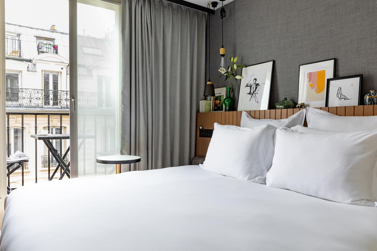 Habitación de hotel con una cama grande, una mesa redonda pequeña, un balcón con vistas a edificios y cuadros en la pared.