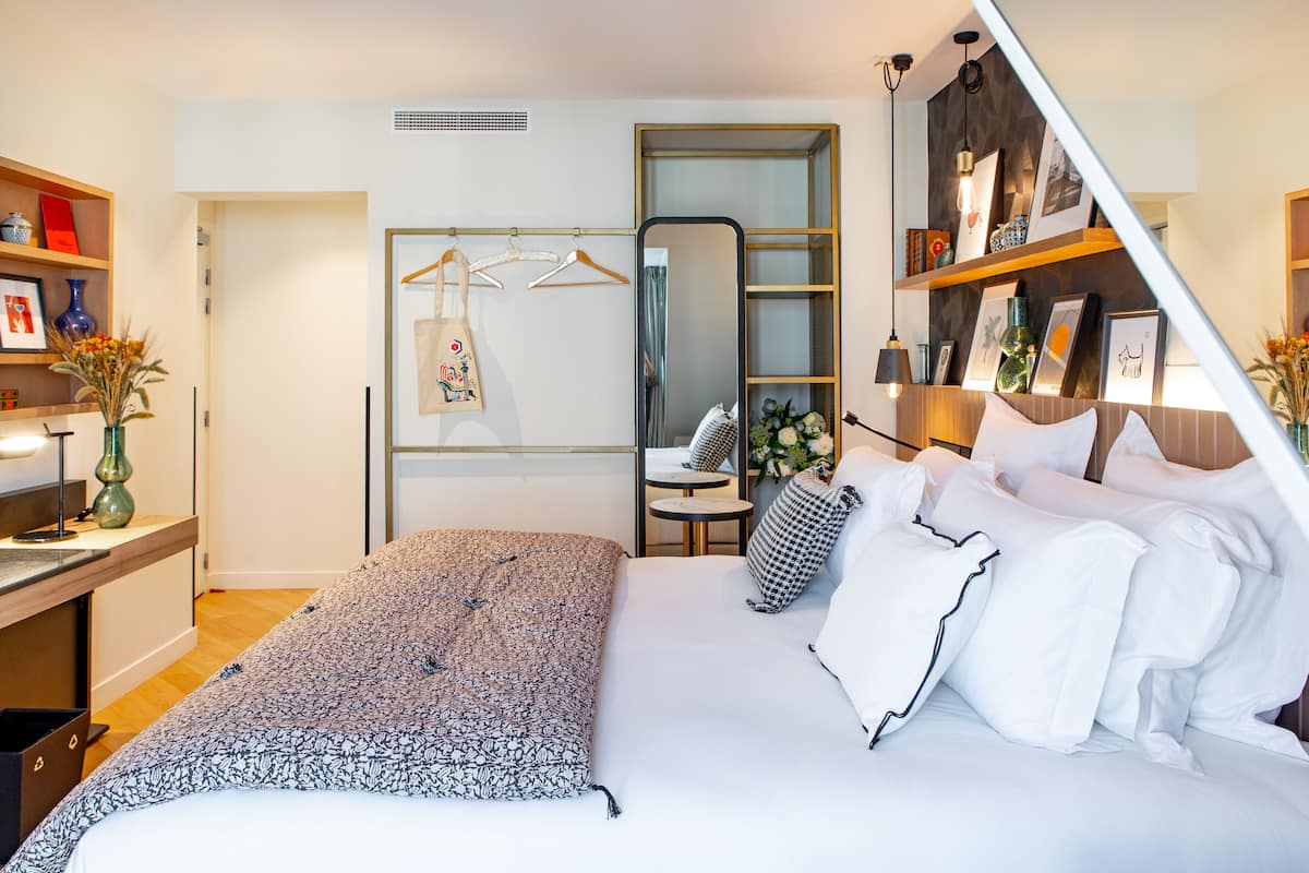 Una habitación de hotel moderna con cama, mesita de noche y espejo. Incluye una pequeña mesa redonda con una silla, una estantería con objetos decorativos y una lámpara colgante.