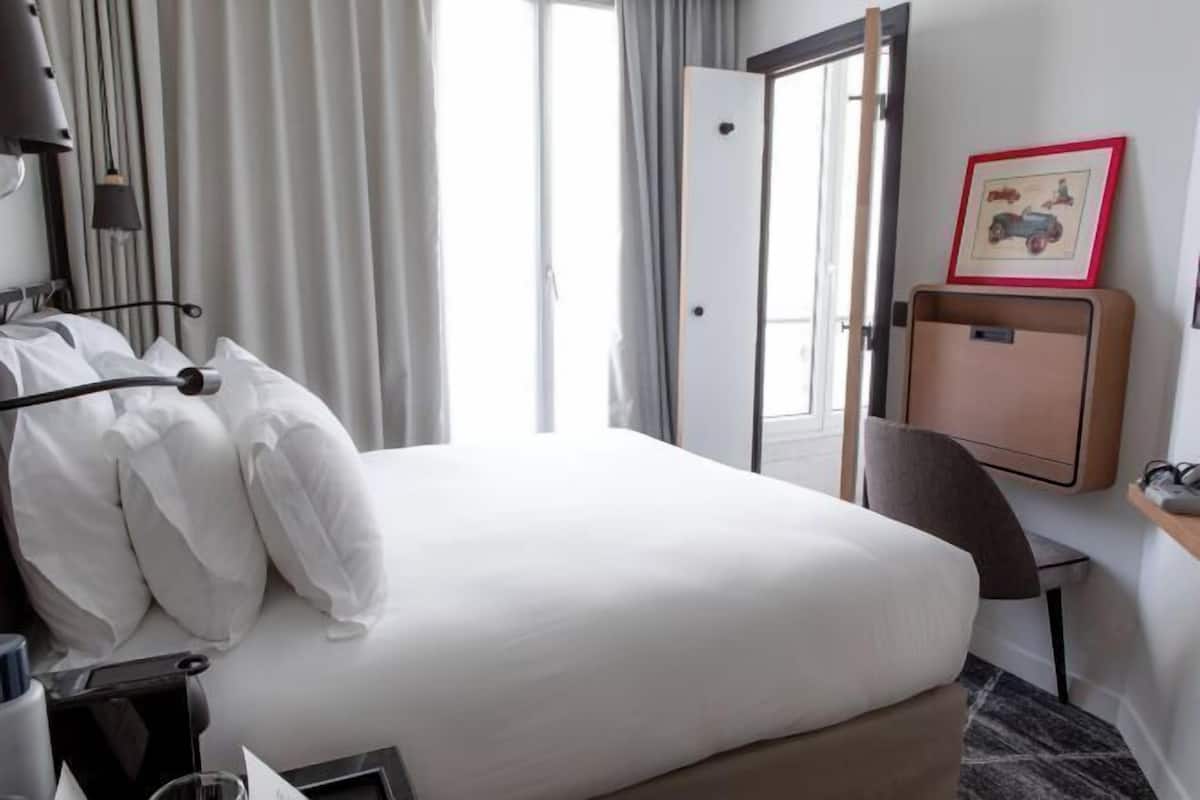 Habitación de hotel con cama, escritorio, silla, televisión y puerta que da a un balcón.