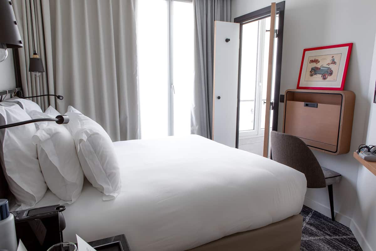 Una habitación de hotel con cama, escritorio, silla, lámpara de escritorio, un marco de fotos y una ventana con cortinas.
