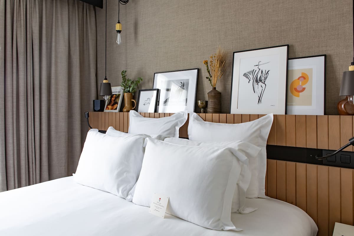 Una cama bien hecha con sábanas y almohadas blancas, un cabecero de madera, y una pared decorada con obras de arte enmarcadas y un jarrón con flores secas.