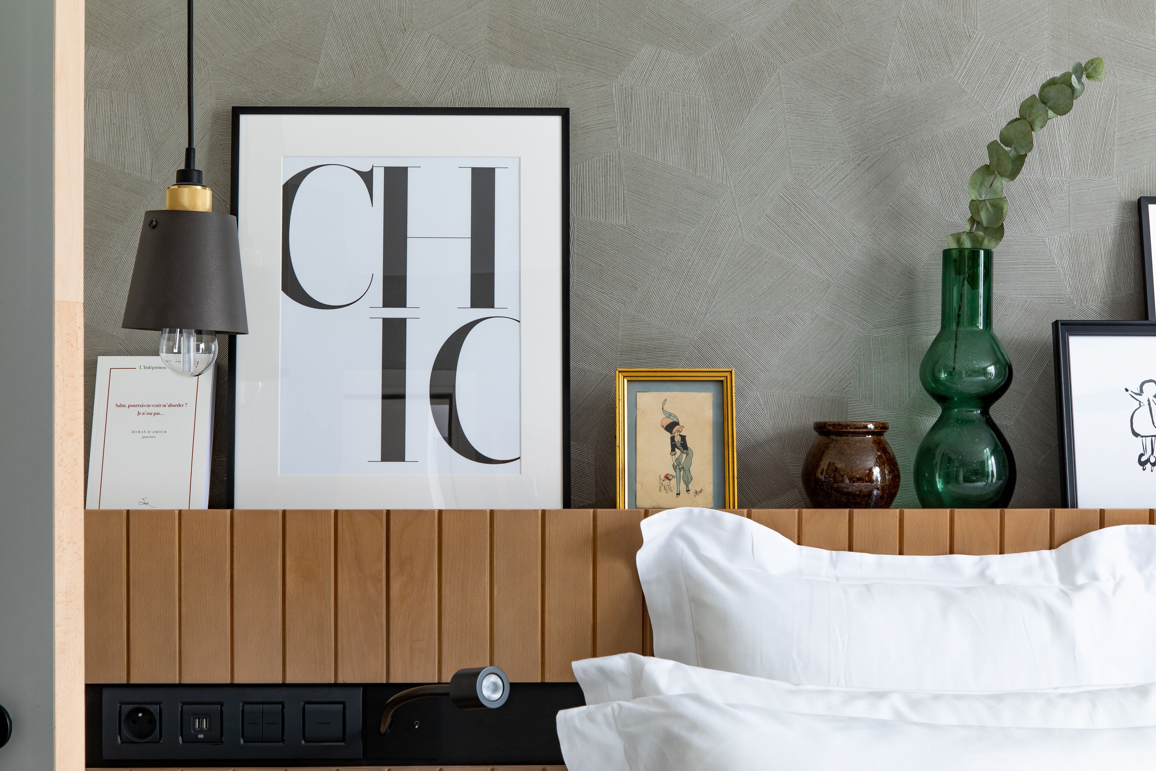A neatly arranged hotel room with a framed 'CHIC' poster, a green vase with a plant, and a wooden headboard.