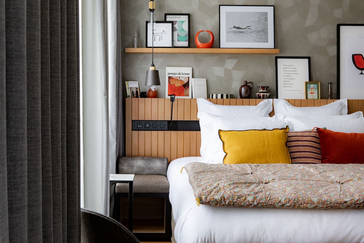 Habitación de hotel con cama, mesita de noche, una silla, estante con fotos enmarcadas y ventana con cortinas.