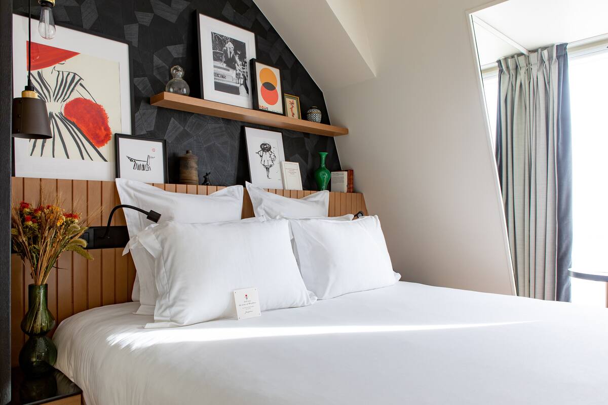 Una cama bien hecha con sábanas blancas, una mesita de noche con un florero y una pared con obras de arte enmarcadas y una estantería con objetos decorativos.