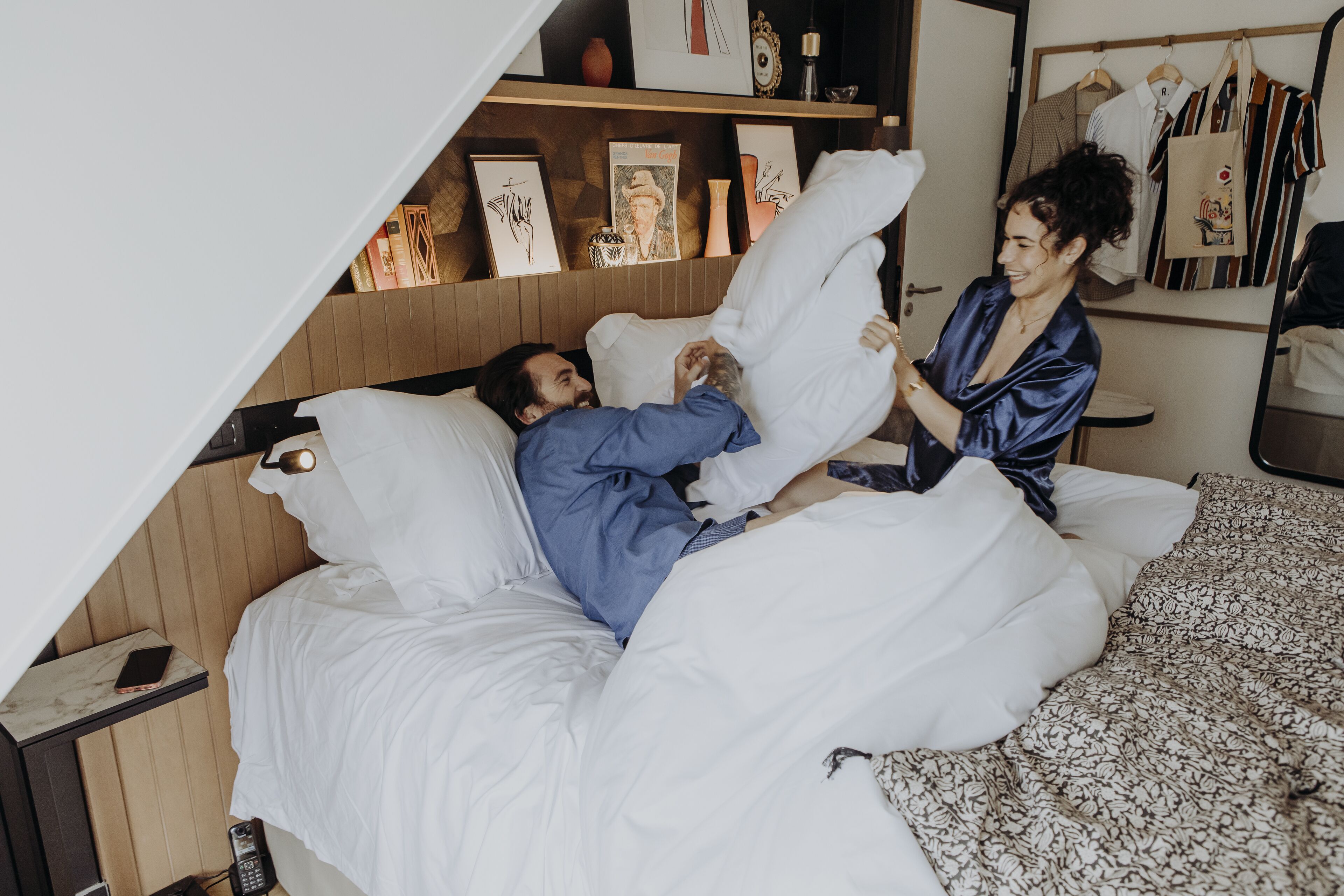 A bedroom with a bed, a bedside table with a phone, and a wall with framed pictures and a mirror.