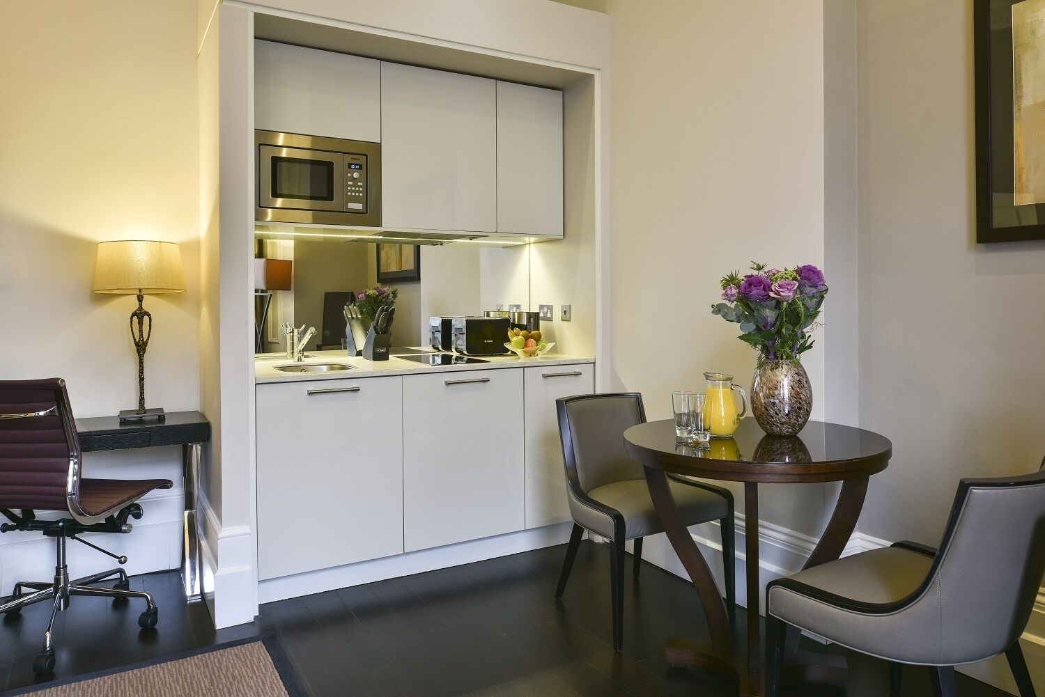 A compact hotel room with a kitchenette, a dining area with two chairs, a small table, and a lamp.